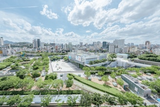 An expansive cityscape with a mix of modern high-rise buildings and lush green parks. The skyline features various architectural styles, with a large open plaza surrounded by numerous trees in the foreground. The sky is dotted with fluffy white clouds under bright sunlight.