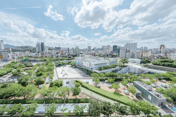 An expansive cityscape with a mix of modern high-rise buildings and lush green parks. The skyline features various architectural styles, with a large open plaza surrounded by numerous trees in the foreground. The sky is dotted with fluffy white clouds under bright sunlight.