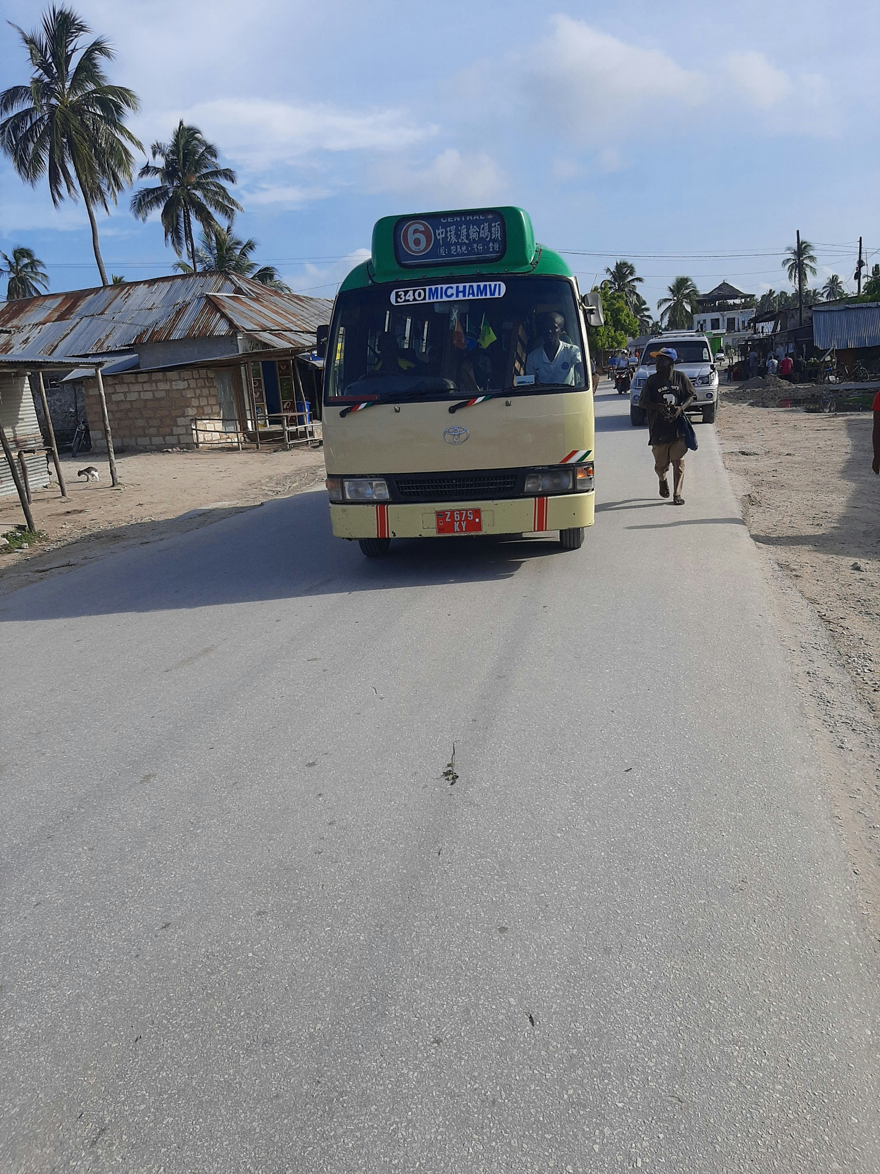 A small bus with a green sign on top indicating route number and destination is traveling down a paved road. Palm trees and simple structures with corrugated roofs line the sides of the road. A person is walking along the road carrying items, and another vehicle follows in the distance. The sky is partly cloudy.