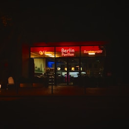 An illuminated cafe and restaurant with a neon sign reading 'Berlin Pavillon'. The establishment is seen at night, with a warm glow emanating from the interior lighting. The street outside appears quiet, and the facade features large glass windows.