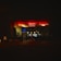 An illuminated cafe and restaurant with a neon sign reading 'Berlin Pavillon'. The establishment is seen at night, with a warm glow emanating from the interior lighting. The street outside appears quiet, and the facade features large glass windows.