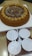 A round cake with a decorative swirl pattern of chocolate and cream on top, surrounded by small round cookies. It is placed on a white cardboard base. Below the cake are four white disposable plates stacked together, accompanied by three plastic spoons.