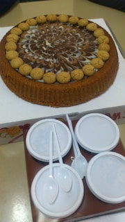 A round cake with a decorative swirl pattern of chocolate and cream on top, surrounded by small round cookies. It is placed on a white cardboard base. Below the cake are four white disposable plates stacked together, accompanied by three plastic spoons.
