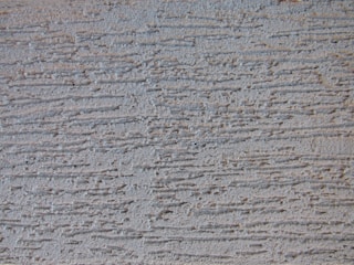 A textured surface with horizontal grooves and ridges, displaying a pattern that resembles rough plaster or stucco. The surface appears to be made of a material like concrete or stone.