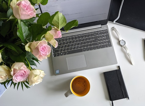 A workspace on a white table featuring an open laptop displaying an email, a bouquet of pink and white roses, a yellow coffee mug, a white watch, and a black notebook with a pen. The setting is well-lit and organized.