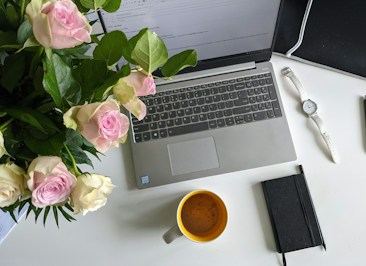 A workspace on a white table featuring an open laptop displaying an email, a bouquet of pink and white roses, a yellow coffee mug, a white watch, and a black notebook with a pen. The setting is well-lit and organized.