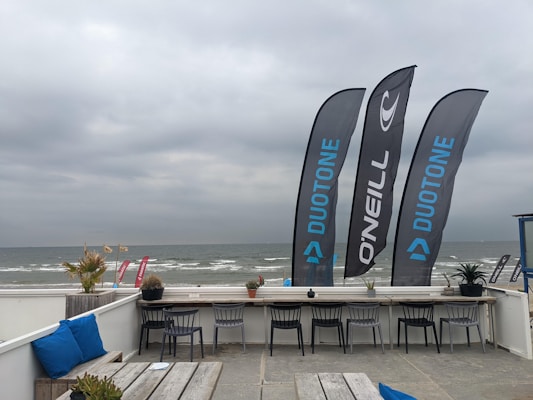 A beachside setting with a gray, overcast sky. Three large flags stand prominently with brand logos, next to a row of chairs lined up at a wooden counter. The ocean waves are visible in the background, and several potted plants are scattered around the area.