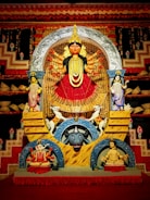 A vividly colored, ornate statue of a multi-armed deity, likely representing a Hindu goddess, is in the center, surrounded by additional smaller figures. The backdrop is elaborately decorated with red and gold patterns, and two animal figures are positioned beneath the goddess, including a buffalo and an elephant.