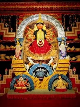 A vividly colored, ornate statue of a multi-armed deity, likely representing a Hindu goddess, is in the center, surrounded by additional smaller figures. The backdrop is elaborately decorated with red and gold patterns, and two animal figures are positioned beneath the goddess, including a buffalo and an elephant.