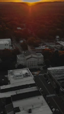 An aerial view of a town at sunset with a prominent courthouse-like building surrounded by other structures. The streets are empty, and trees are visible in the background.