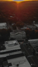 An aerial view of a town at sunset with a prominent courthouse-like building surrounded by other structures. The streets are empty, and trees are visible in the background.
