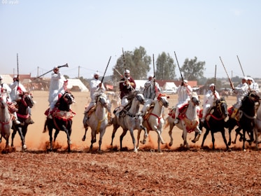 A group of traditionally dressed riders is performing a cavalry display on horseback across a dusty field. The riders are wearing white robes and holding rifles in the air. Their horses are adorned with colorful harnesses.