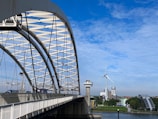 A large steel arch bridge extends over a river, with vehicles traveling on it. In the background, a wind turbine is visible against a bright blue sky with scattered clouds. Buildings, including a tall white structure and a modern glass one, line the riverbank surrounded by greenery.