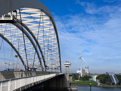 A large steel arch bridge extends over a river, with vehicles traveling on it. In the background, a wind turbine is visible against a bright blue sky with scattered clouds. Buildings, including a tall white structure and a modern glass one, line the riverbank surrounded by greenery.