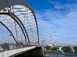 A large steel arch bridge extends over a river, with vehicles traveling on it. In the background, a wind turbine is visible against a bright blue sky with scattered clouds. Buildings, including a tall white structure and a modern glass one, line the riverbank surrounded by greenery.