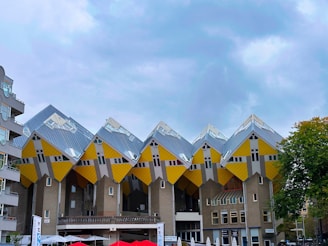 A row of uniquely designed cube-shaped buildings stands against a cloudy sky. The structures feature slanted walls and roofs, with a bright yellow and gray color scheme. Surrounding buildings with more traditional designs are visible, and there are outdoor seating areas with red umbrellas in the foreground.
