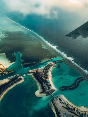 Aerial view of a coastal area with turquoise waters and scattered islands. Overwater bungalows are visible, arranged in curved patterns extending into the lagoon. The wing of an airplane can be seen in the top right corner, adding perspective to the scene. Waves gently lap along the coral reef edge, and patches of vegetation can be seen on the sandy islands.