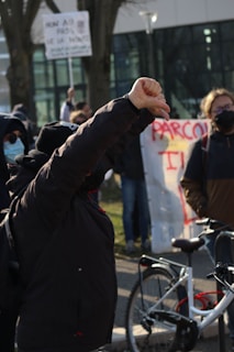 A group of people appears to be participating in a protest. One individual in the foreground raises their fist with determination. They are wearing a dark jacket and a beanie. Some members of the group are wearing masks, and various signs with text are visible, though not entirely legible. A bicycle is partially visible on the right side.