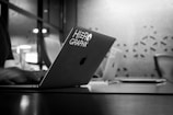 A black and white image showing a laptop with 'HERO GRAPHX' sticker in a modern office setting. The focus is on the laptop, while the background is softly blurred, including a table, chairs, and patterned wall.