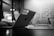 A black and white image showing a laptop with 'HERO GRAPHX' sticker in a modern office setting. The focus is on the laptop, while the background is softly blurred, including a table, chairs, and patterned wall.