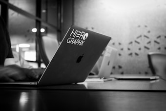 A black and white image showing a laptop with 'HERO GRAPHX' sticker in a modern office setting. The focus is on the laptop, while the background is softly blurred, including a table, chairs, and patterned wall.