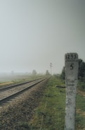 A railway track stretches into the distance alongside a lush green field, with a weathered stone milestone marked with the numbers 1295 and 5 in the foreground. The sky appears overcast, and there are blurred trees and a signal post visible in the background.
