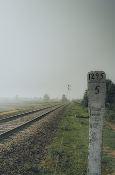 A railway track stretches into the distance alongside a lush green field, with a weathered stone milestone marked with the numbers 1295 and 5 in the foreground. The sky appears overcast, and there are blurred trees and a signal post visible in the background.