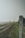 A railway track stretches into the distance alongside a lush green field, with a weathered stone milestone marked with the numbers 1295 and 5 in the foreground. The sky appears overcast, and there are blurred trees and a signal post visible in the background.