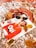 A magazine cover featuring a person wearing a red sweater with the word 'Oui' is placed on a patterned fabric. Surrounding the magazine are autumn leaves, a pair of sunglasses, a lipstick, and a small perfume bottle, creating a cozy, fall-themed aesthetic.
