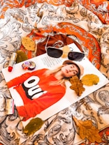 A magazine cover featuring a person wearing a red sweater with the word 'Oui' is placed on a patterned fabric. Surrounding the magazine are autumn leaves, a pair of sunglasses, a lipstick, and a small perfume bottle, creating a cozy, fall-themed aesthetic.