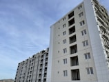 A series of modern multi-story apartment buildings under a clear blue sky. The buildings are tall with multiple windows, and one of the structures appears to be under construction with scaffolding visible.