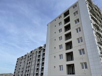 A series of modern multi-story apartment buildings under a clear blue sky. The buildings are tall with multiple windows, and one of the structures appears to be under construction with scaffolding visible.