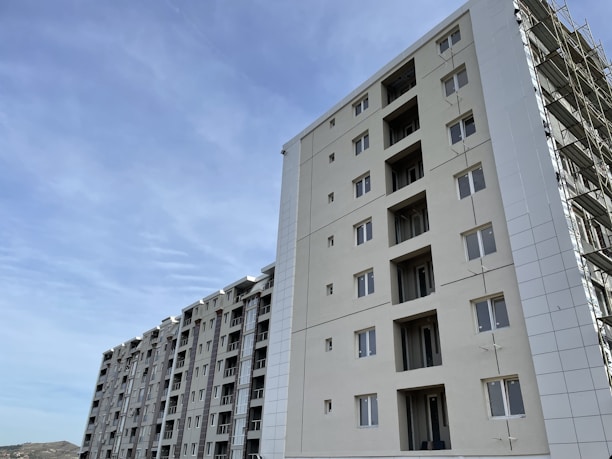 A series of modern multi-story apartment buildings under a clear blue sky. The buildings are tall with multiple windows, and one of the structures appears to be under construction with scaffolding visible.