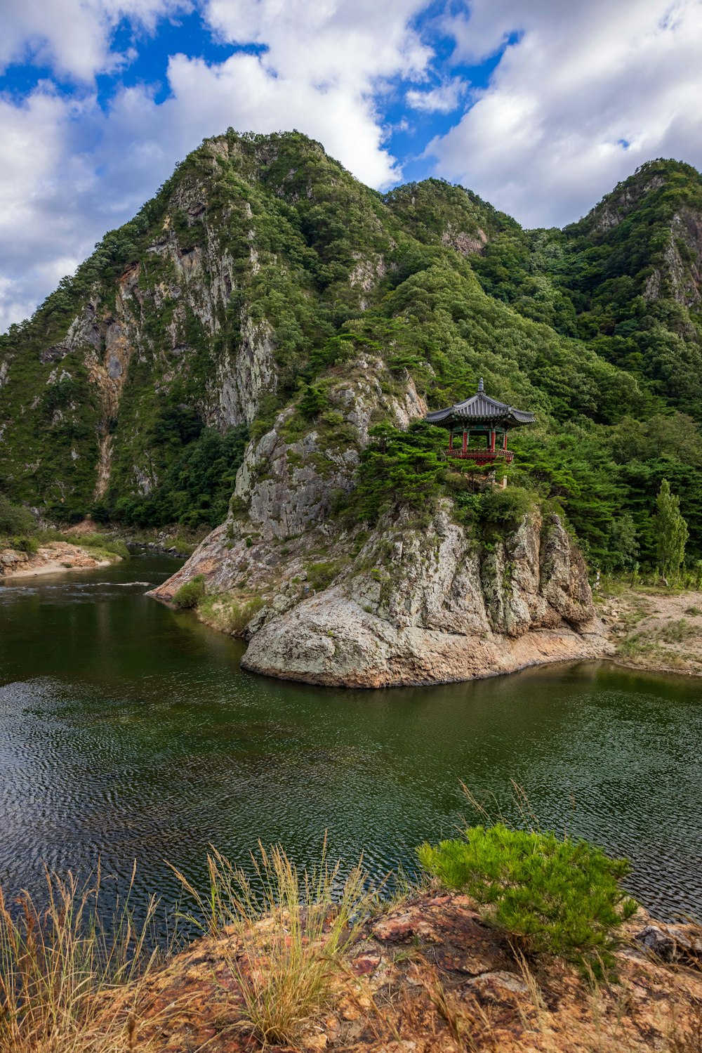 a building on a rock in the water with trees and mountains in the background