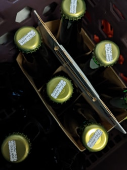 Several glass bottles are positioned upright in a cardboard carrier, with some parts of a dark plastic crate visible around them. The caps of the bottles are green in color with small printed text on them.