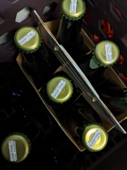 Several glass bottles are positioned upright in a cardboard carrier, with some parts of a dark plastic crate visible around them. The caps of the bottles are green in color with small printed text on them.