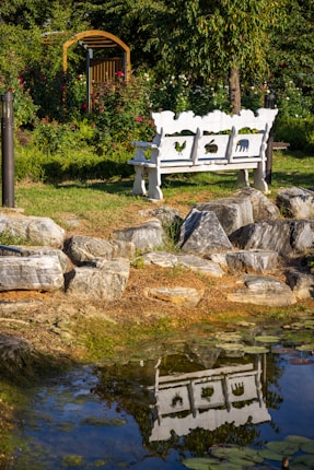 A serene garden scene featuring a white decorative bench with cut-out animal shapes located near a rock-lined pond. The bench is reflected in the still water, surrounded by lush green foliage and vibrant flowers. In the background, there is a wooden arbor and a lush garden with trees and a variety of plants.