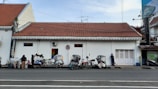 A row of becak (pedicabs) parked in front of a white building with a red tiled roof. The building has small colorful windows and a no-parking sign. A few people are interacting; one is standing near a cart selling goods.