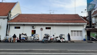 A row of becak (pedicabs) parked in front of a white building with a red tiled roof. The building has small colorful windows and a no-parking sign. A few people are interacting; one is standing near a cart selling goods.