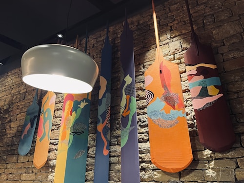 Several artistically painted paddles hang vertically against a textured brick wall. Each paddle features a unique design with abstract patterns and vibrant colors, creating a striking visual effect. A large white dome-shaped light fixture is suspended from the ceiling, casting light over the scene.