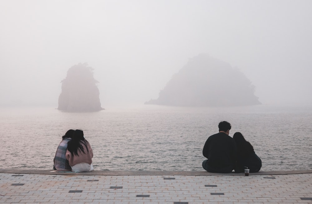 a man and a woman sitting on a stone walkway looking at a large rock in the water