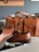 A brown leather briefcase and a pair of brown leather dress shoes are arranged on a light-colored surface. The briefcase features large pockets with flap enclosures, buckles, and stitching details. The shoes have a polished finish with visible laces. In the background, there's a wall with a framed picture.