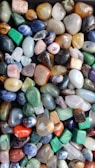 A collection of polished gemstones in various shapes and sizes, displaying a wide range of vibrant colors. The stones have a smooth surface, reflecting light and highlighting their natural patterns and textures.