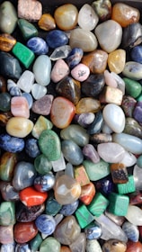 A collection of polished gemstones in various shapes and sizes, displaying a wide range of vibrant colors. The stones have a smooth surface, reflecting light and highlighting their natural patterns and textures.