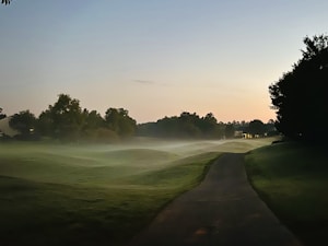 A serene landscape featuring a misty golf course during sunrise or sunset. A narrow paved path winds through the lush green grass, flanked by groups of trees. Soft mist gently hovers over the undulating terrain, creating a tranquil and ethereal atmosphere.