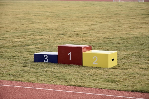 podium avec la marche 3 bleue, la marche 2 jaune et la marche 1 rouge posé sur l'herbe au bord d'une piste de course rouge