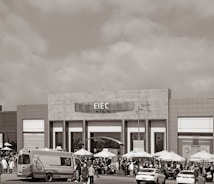 A large building with signage reading 'EIEC' is visible, surrounded by a crowd of people gathered in front of it. Vehicles, including a van and a car, are parked nearby. Several umbrellas are set up outside the building, possibly for outdoor seating or shelter.