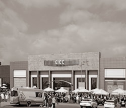 A large building with signage reading 'EIEC' is visible, surrounded by a crowd of people gathered in front of it. Vehicles, including a van and a car, are parked nearby. Several umbrellas are set up outside the building, possibly for outdoor seating or shelter.
