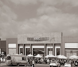 A large building with signage reading 'EIEC' is visible, surrounded by a crowd of people gathered in front of it. Vehicles, including a van and a car, are parked nearby. Several umbrellas are set up outside the building, possibly for outdoor seating or shelter.