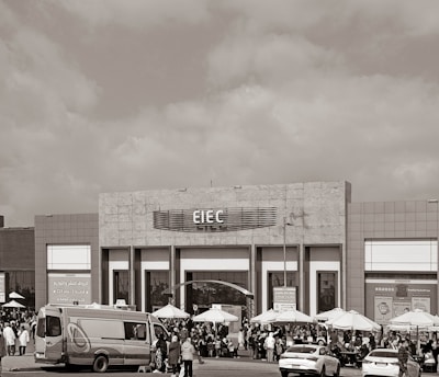 A large building with signage reading 'EIEC' is visible, surrounded by a crowd of people gathered in front of it. Vehicles, including a van and a car, are parked nearby. Several umbrellas are set up outside the building, possibly for outdoor seating or shelter.
